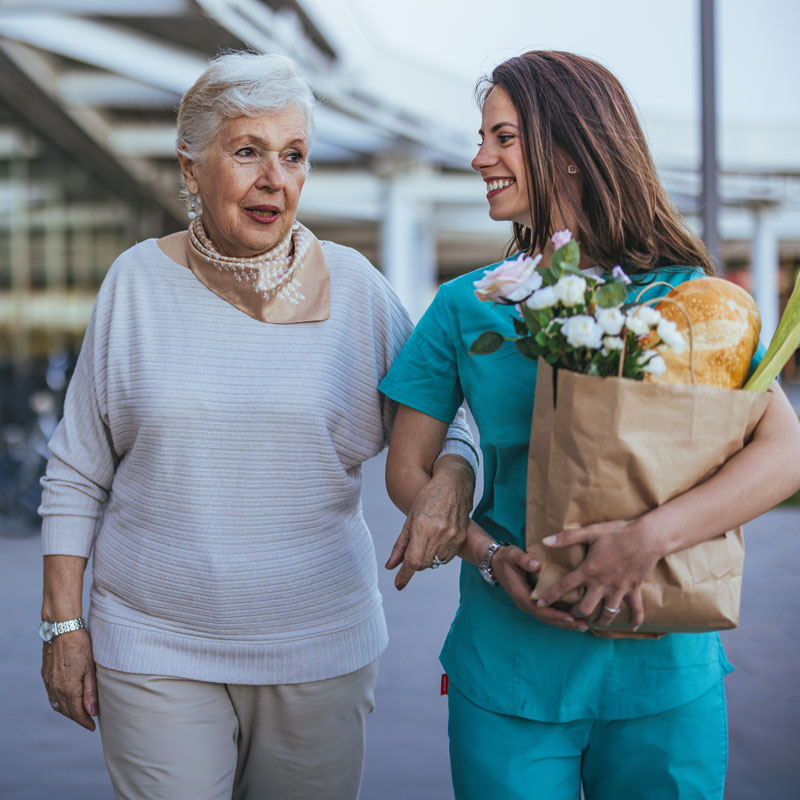Ein junge Frau mit einer Einkaufstasche in der Hand hält eine ältere Dame am Arm und begleitet sie.